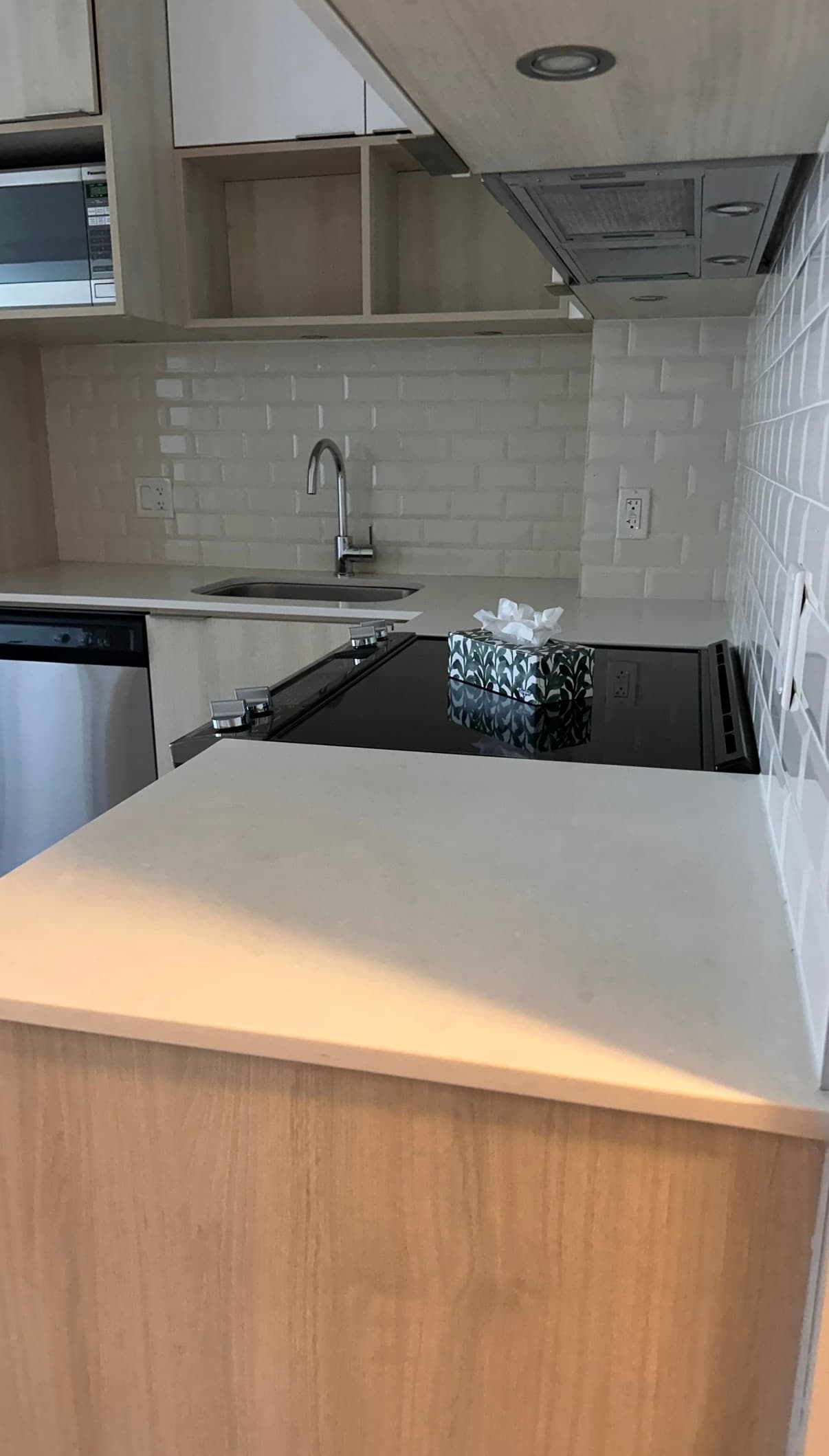 Modern kitchen with light wood cabinets, white subway tile, stainless sink, and black glass cooktop.