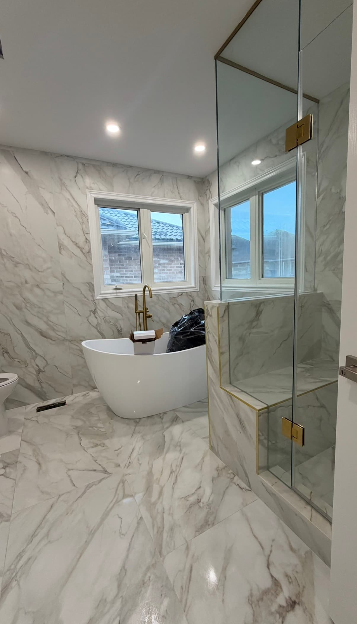 Modern marble bathroom featuring a white freestanding tub, gold fixtures, and a glass-enclosed shower.