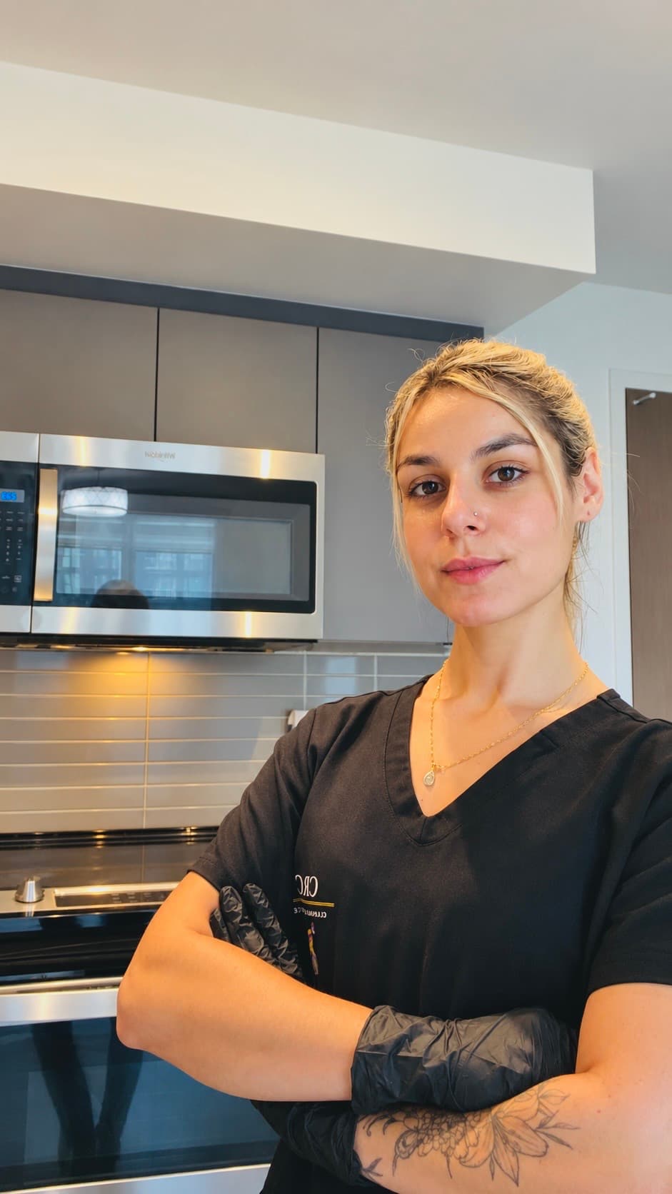 Blonde woman in black scrubs and gloves stands with arms crossed in a modern kitchen.